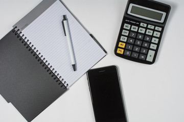 Flat lay of calculator, phone and notebook with a pen on a white table.