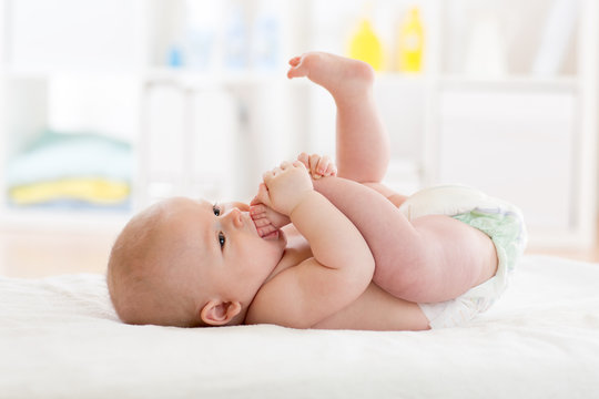 Funny Little Baby Wearing A Nappy Playing With His Legs In Nursery. Cute Kid Sucking His Feet. Child After Bath Or Shower On Bed. Infant Skin Care.