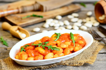 Homemade beans stew on a plate and on old wooden table. White beans stewed with carrots and tomato sauce and served with green arugula. Delicious and hearty beans side dish. Rustic style. Closeup