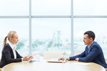 Profile view of two business people, man and woman,   representing sides of contract, sitting opposite each other at table discussing deal during meeting in office