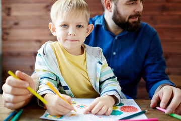 Cute little blond-haired boy with deep grey eyes sitting on laps of his bearded father, looking at camera and holding pencil in hand for coloring picture