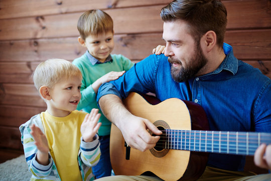 Bearded nursery teacher giving music lesson to little boys: he playing guitar and singing funny song while his pupils listening to him with interest
