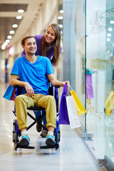 Fototapeta premium Handicapped young man in wheelchair having good time with his loving girlfriend in mall, walking with shopping bags and smiling cheerfully