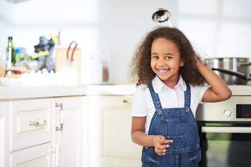 Cute little girl in denim jumpsuit standing next to kitchen oven and looking at camera with toothy smile, waist-up portrait