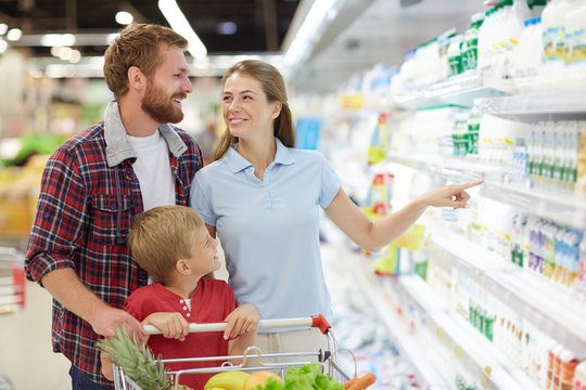 Family of three choosing goods in hypermarket: pretty woman pointing at shelf with dairy products while her family looking at it with toothy smiles