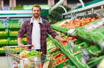 Portrait of young bearded man in checked shirt stretching arm towards tomato while looking at camera with toothy smile in supermarket