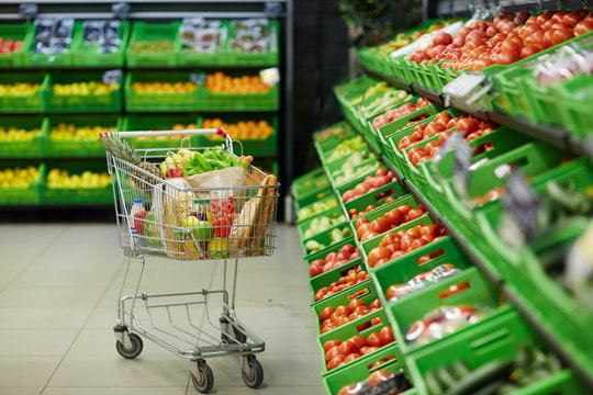 Metal shopping trolley with healthy grocery items standing in fruit and vegetable department of hypermarket