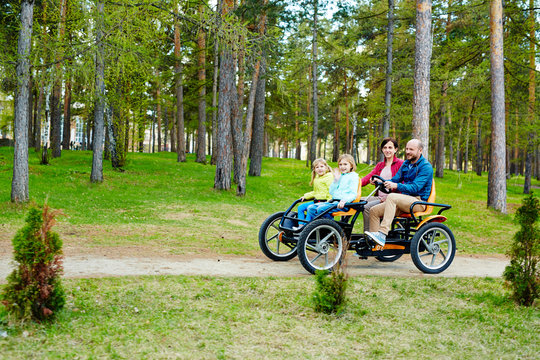 Cheerful family of four riding orange quadricycle pedal car while enjoying spring in park with pine forests