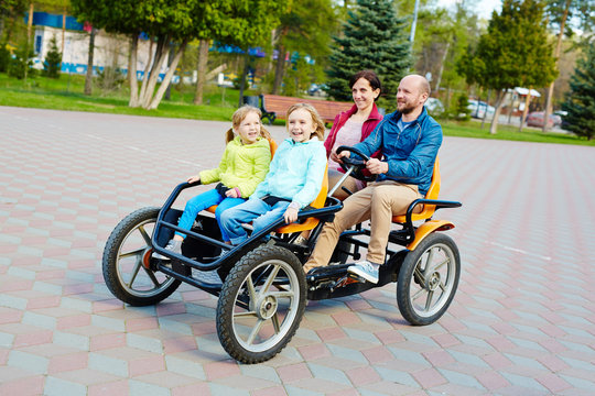 Pretty family having fun in spring park: bearded middle-aged father driving orange quadricycle pedal car while his joyful family sitting in it with wide smiles