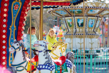 Two blond-haired little girls in bright windbreakers sitting on carousel horses and smiling happily