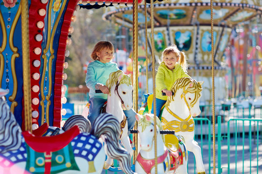 Cute little sisters enjoying spring in funfair: they riding on colorful carousel and looking at camera with wide smiles