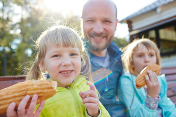Blond-haired little girls sitting on wooden park bench with their bald dad and eating tasty hotdogs, waist-up portrait