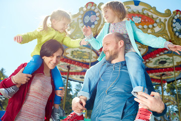 Carefree little girls playing with each other while sitting on shoulders of their smiling middle-aged parents, colorful merry-go-round observed on background © pressmaster
