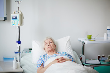 Sick elderly woman resting in hospital bed after getting drop bottle treatment, looking melancholy...