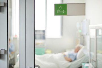 Hospital ward sign on closed glass door in clinic with blurred shape of senior patient lying on bed...
