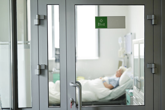 Wide shot image of glass door in clinic with green ward sticker on it closed with key, locking senior patient sleeping on bed inside