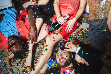 Tipsy multiethnic group of friends relaxing on floor with colorful confetti while celebrating holiday together