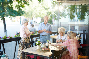 Four concentrated senior friends gathered in lovely outdoor cafe and playing cards, tasty cheesecake standing on corner of table