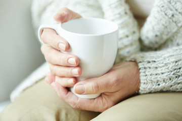 Elderly woman in warm home clothing sitting on couch and holding white cup of coffee in hands,...