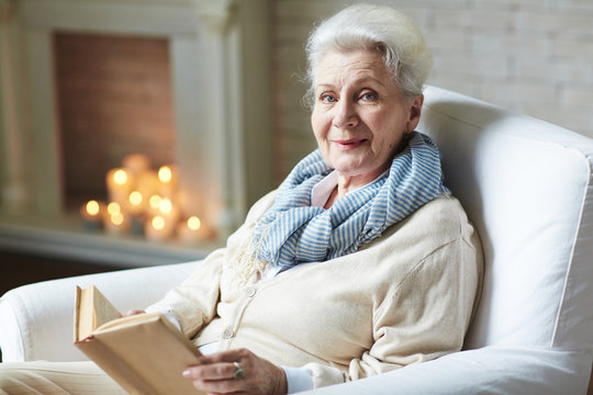 Portrait of smiling attractive retired woman in beige cardigan distracted for second from reading book in order to pose for photography