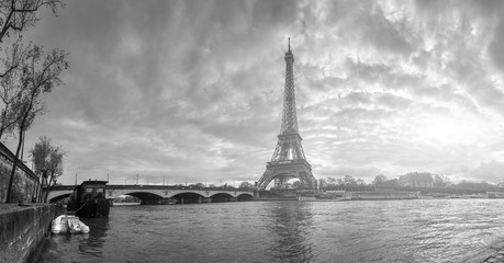Beautiful panoramic view of the Eiffel Tower and Jena bridge from the river Seine embankment. Dramatic cloudscape. Traditional sitycape in backlit morning sunbeam. BW photography. Paris, France.