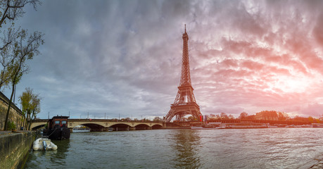 Beautiful panoramic view of the Eiffel Tower and Jena bridge from the river Seine embankment. Dramatic cloudscape. Traditional citycape in backlit morning sunbeam. Paris, France.