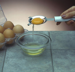 hand holding an egg separator with yolk inside and the white falling into a bowl, a sixpack of eggs in the background