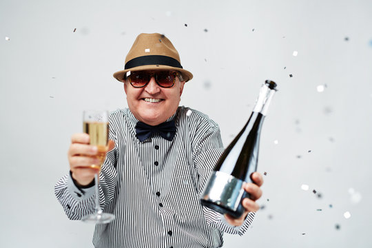 Waist-up Portrait Of Excited Elderly Man In Bowler Hat And Sunglasses Cheering With Bottle Of Champagne And Confetti