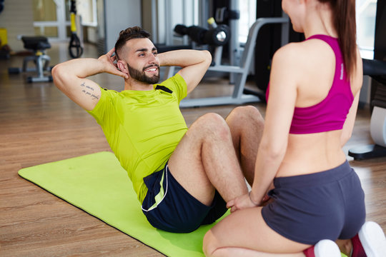 Working out in gym: young girl holding legs of her partner while he is doing sit ups, both wearing bright sportswear