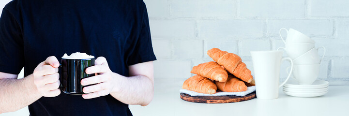 Black mug with coffee drink, cream topping in hands of young man in black/ddark blue t-shirt, copy space. Wide panoramic image. White kitchen background with pastry and dishes.