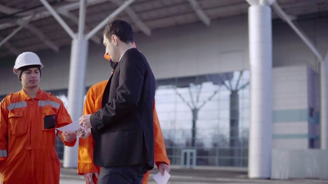 Manager in a suit giving envelopes with money to workers in orange uniform and helmets. Successful finish of the project. Shot in 4K