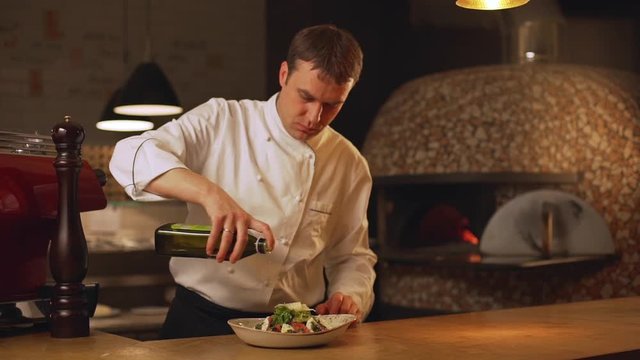Confident Caucasian chief cook pouring olive oil from bottle on tomato and cheese salad in soup plate on wooden table in slowmotion