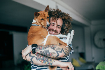 Crazy haired hipster in striped shirt and tattoos hugging dog in industrial loft apartment