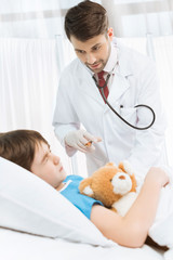 Scared little boy with teddy bear lying in hospital bed while doctor making injection