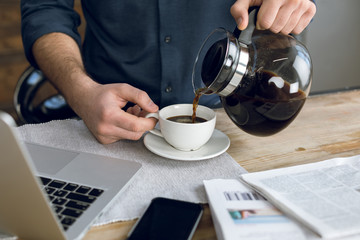 Partial view of man pouring coffee in cup on desk with laptop and smartphone