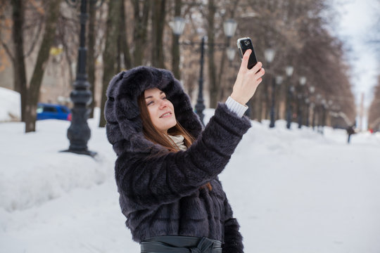 young beautiful girl in a fur coat on a walk in a winter park with a telephone in hands