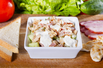 Simple, fresh salad with vegetables and meat in white bowl on a wooden board next to toast, grilled chicken and smoked ham.