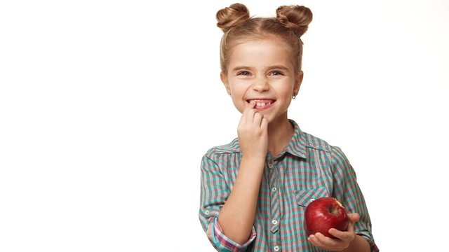 Small cute kid Caucasian girl in plaid shirt and two hair buns standing on white background biting red apple and touching teeth in slowmotion