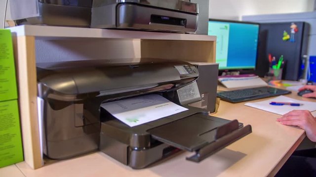 A worker in the office is printing something out of his printer. It is a colourful print. Then he takes the paper and looks at it.
