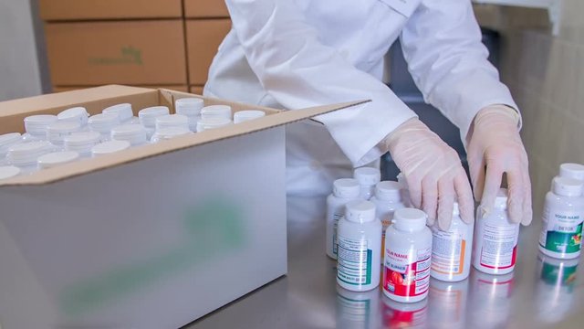 A pharmaceutical factory worker is organizing small white cars of the same size into a big box. A few of them can't fit inside because the box is already full and they are standing on a table.

