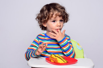 Kid getting messy while eating a chocolate cake. Beautiful curly hair boy eating sweets. Toddler in high chair being hungry.