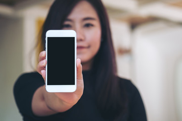 Mockup image of a beautiful woman holding and showing white mobile phone with blank black screen and smiley face in vintage cafe