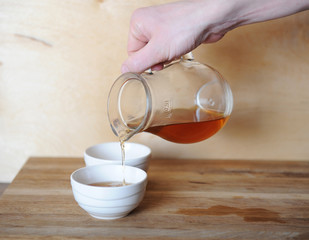 Hand pouring tea or coffee from a glass decanter on the porcelain bowl. Wooden background.