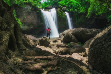 Young man standing interesting in front of waterfall and take photo by smartphone, beautiful waterfall in thailand, soft focus and blur due to long exposure.