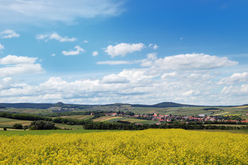 Rural landscape with a cozy village among the hills, fields and meadows under cloudy blue sky
