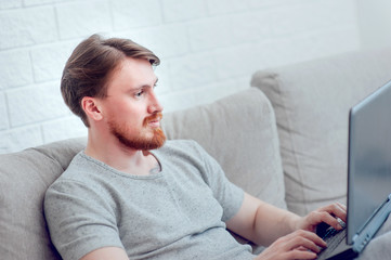 A bearded man with a laptop sitting on the sofa at home