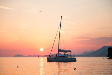 Yacht is sailing with people on background of sunset, sea, mountains, sky, sun.
