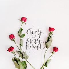 Inspirational quote "Enjoy Every Moment" written in calligraphic style on paper with red roses isolated on white background. Flat lay, top view