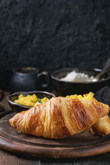 Breakfast with two croissant, butter, cottage cheese, coffee and sliced mango fruit, served on wood chopping board over old wooden background. Close up.