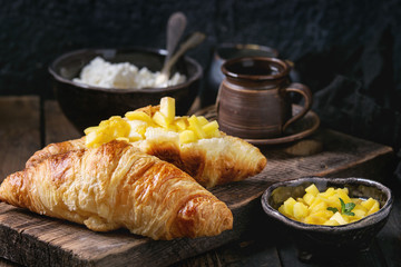 Breakfast with two croissant, butter, cottage cheese, coffee and sliced mango fruit, served on wood chopping board over old wooden background. Close up.